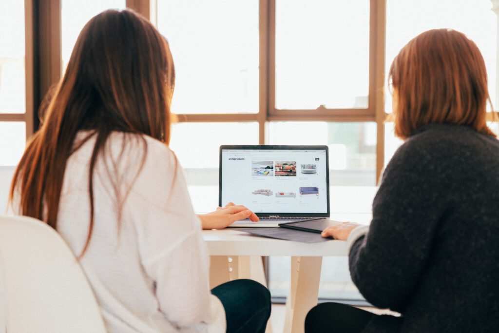 Zwei Frauen sitzen vor einem Laptop. Eine davon bedient den Laptop mit einem Touchscreen.