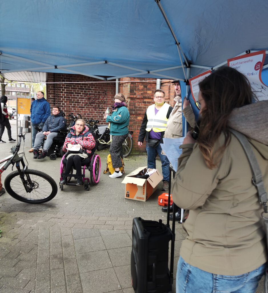 Protest des ZSL Nord vor dem Husumer Bahnhof. Mehrere Personen sind zu sehen, teilweise im Rollstuhl. Im Vordergrund steht eine EUTB-Beraterin in einem Pavillon mit einem Mikrofon, die eine Protestrede hält.