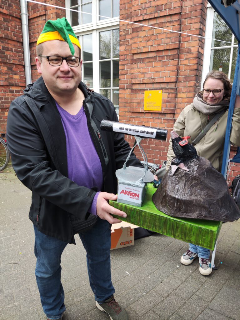 Zwei EUTB-Beratende beim Protest vor dem Husumer Bahnhof. Eine Person trägt eine Kasperlmütze auf dem Kopf und hält einen selbstgebastelten Preis für die Deutsche Bahn in der Hand. Dieser prangert die mangelnde Barrierefreiheit an dem Bahnhof an.