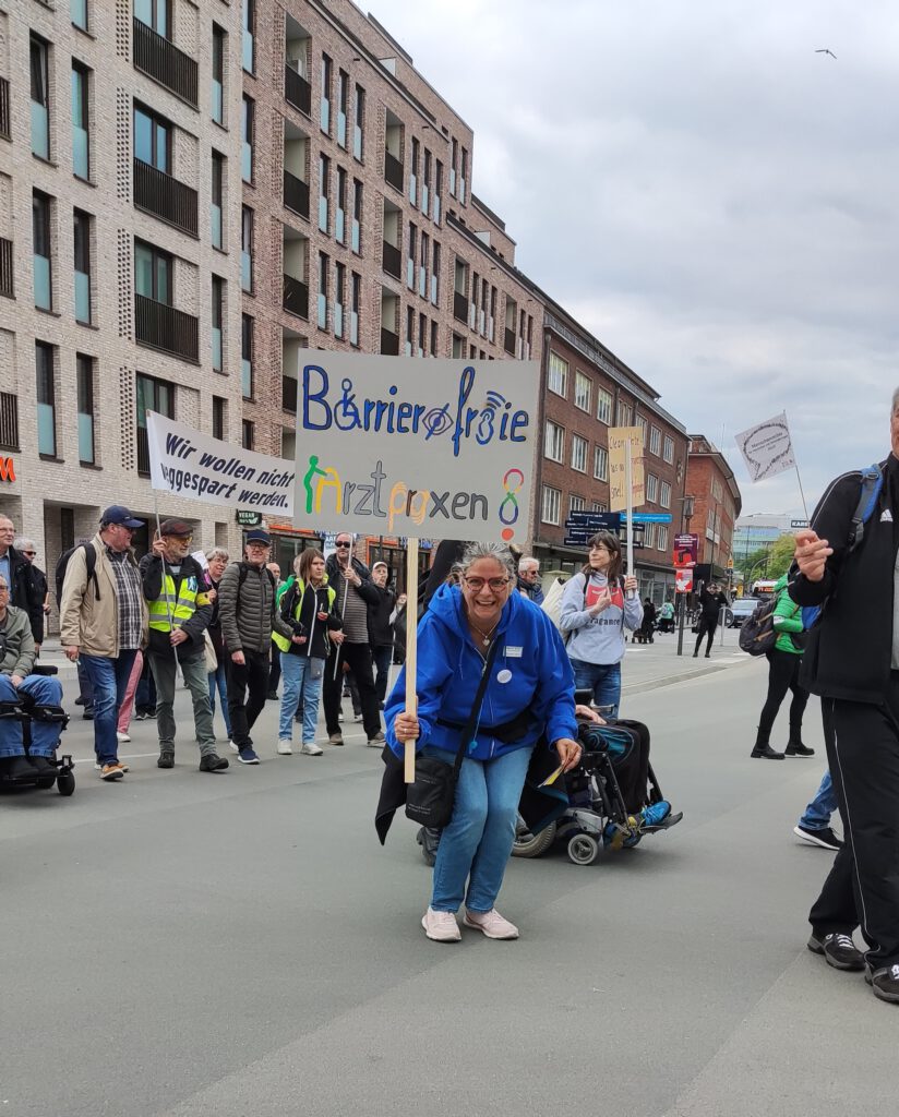 Im Vordergrund eine Frau mit einem Protestschild auf dem steht: "Barrierefreie Arztpraxen". Im Hintergrund ein Demozug