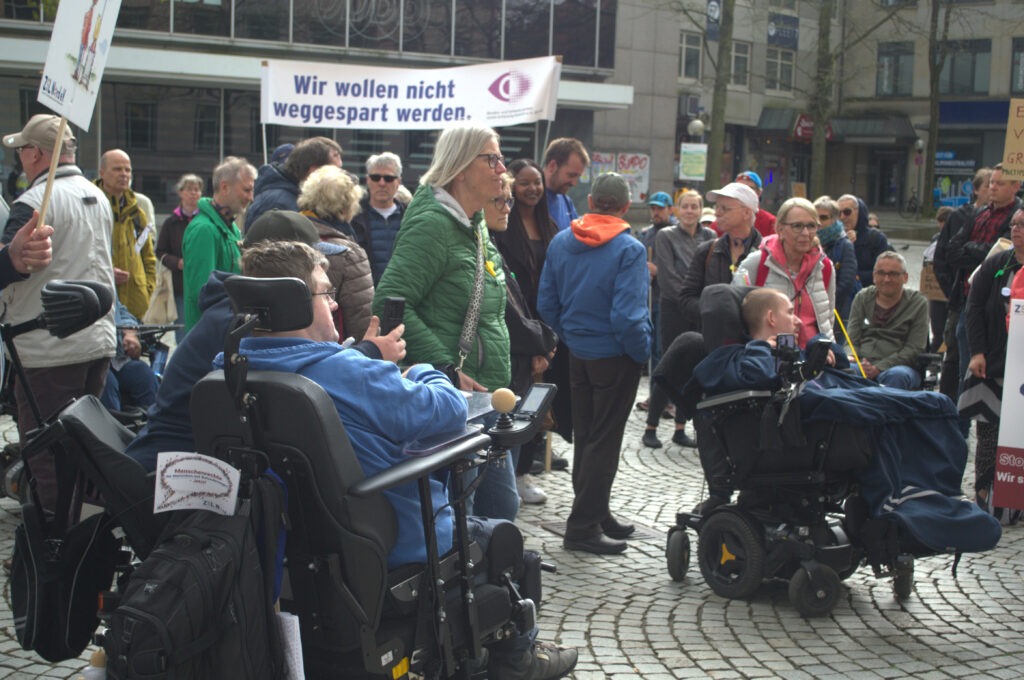 Demonstrierende mit Protestplakaten. Im Vordergrund zwei Menschen im Rollstuhl.