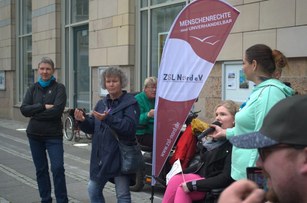 Geschäftsführerin des ZSL Nord, Janine Kolbig, hält eine Rede. Daneben Sprachdolmetscherinnen, die übersetzen und eine ZSL Fahne.